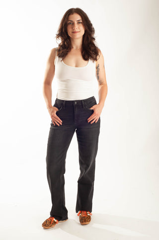 A woman modeling a pair of dark wash straight-leg jeans with a white tank top and patterned sneakers, standing against a plain white studio background.