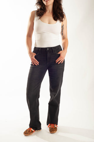 A woman modeling a pair of dark wash straight-leg jeans with a white tank top and patterned shoes, standing against a plain white studio background.