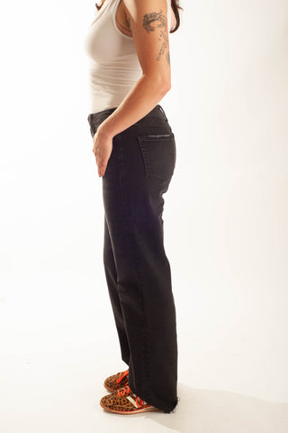 A side-profile view of a person wearing black wide-leg jeans, a white tank top, and patterned slip-on shoes against a plain white studio background.