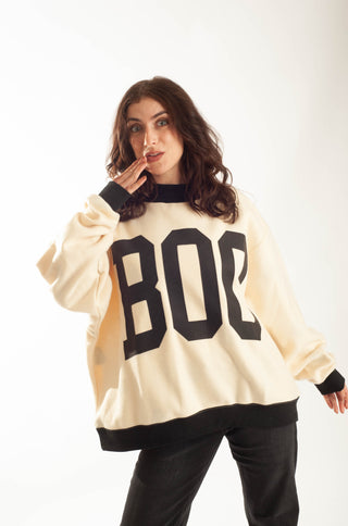 A woman modeling a cream-colored oversized sweatshirt with bold black collegiate-style lettering that reads 'BOB' across the front, paired with black pants, against a plain white studio background.