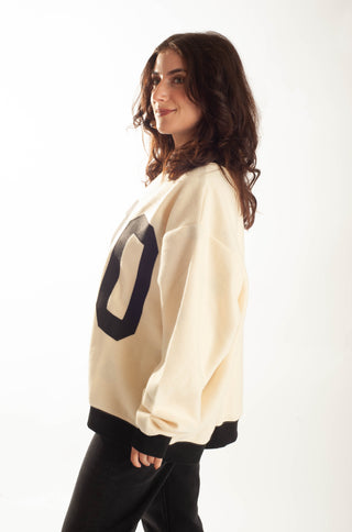 A woman wearing a cream-colored crewneck sweatshirt with a large black number '0' on the back and black ribbed cuffs and hem, shown from a side profile against a white studio background.