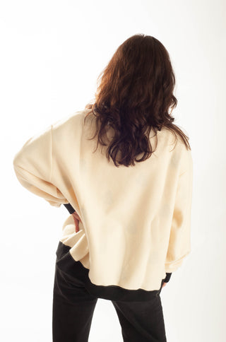A woman with long, wavy brown hair is shown from behind, wearing a cream-colored oversized sweatshirt and black pants against a plain white background.