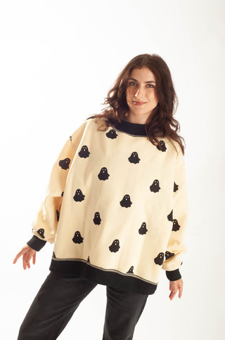 A woman modeling a cream-colored oversized sweatshirt with black ghost patterns and black ribbed trim, paired with black pants, against a plain white background.