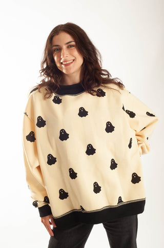 A woman wearing a cream-colored crewneck sweatshirt with a black ghost pattern and black ribbed trim, paired with black pants, against a plain white background.
