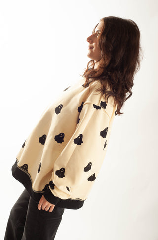 A woman wearing a cream-colored oversized sweatshirt with black ghost patterns and black ribbed cuffs and hem, paired with black pants, photographed against a plain white background.