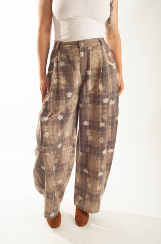 A person wearing loose, wide-leg plaid pants in brown, beige, and white, paired with a white sleeveless top and brown shoes, photographed against a plain white studio background.
