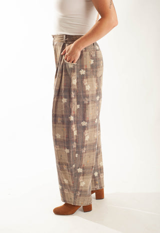 A person shown from the waist down wearing loose, wide-leg plaid pants in beige, brown, and white with small floral motifs, paired with brown heeled boots against a plain white studio background.