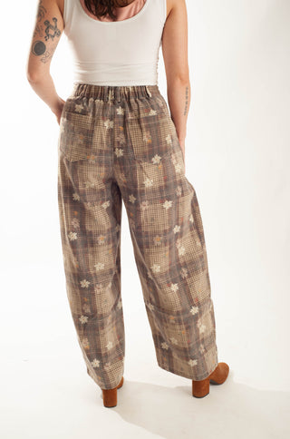 A person wearing loose, wide-leg plaid pants in brown, beige, and cream tones with small floral motifs, paired with a white tank top and brown heels, photographed against a plain light background.