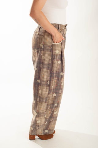 A side view of a person wearing wide-leg plaid trousers in brown, beige, and white, paired with a white top and brown heels, against a plain white studio background.