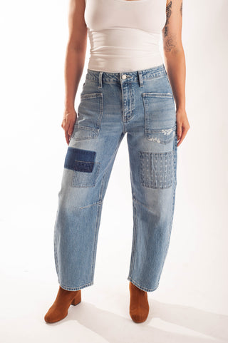 A model wearing light blue patchwork denim jeans with distressed details and brown heeled boots, shown from the waist down against a white studio background.