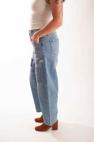 A side-profile view of a person wearing light-wash blue patchwork denim jeans, a white tank top, and brown block-heel boots against a clean white studio background.