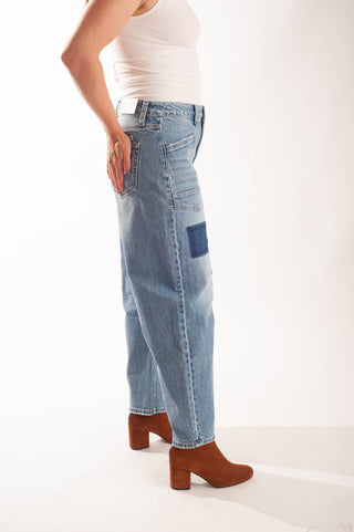 A model wearing light-wash blue denim jeans with a dark blue patch pocket on the side, styled with a white tank top and brown block-heel ankle boots against a white studio background.