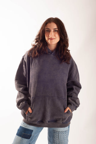 A woman with long brown hair wearing an oversized dark charcoal gray washed hoodie and light blue patchwork jeans, standing against a plain white studio background.