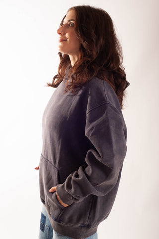 A woman with long brown hair is shown in profile wearing an oversized dark charcoal gray sweatshirt and light blue jeans, standing against a plain white studio background.