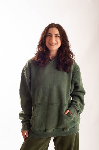 A woman with long wavy brown hair wearing an oversized olive green hoodie and matching green pants, standing against a plain white studio background.