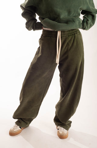A person wearing loose, oversized olive green sweatpants with a cream drawstring, paired with a matching olive green sweatshirt and white sneakers, photographed against a clean white studio background.