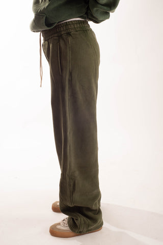 A person wearing loose olive green sweatpants with a drawstring waist, paired with a dark green top and brown sneakers, photographed against a plain white studio background.