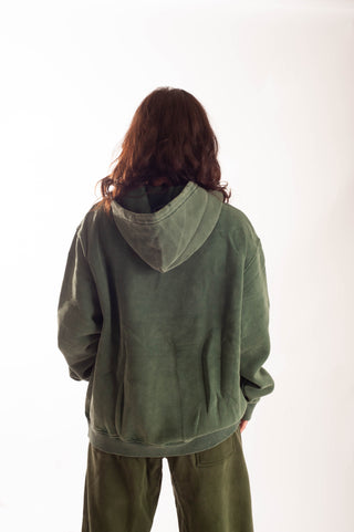 A person viewed from behind wearing an oversized sage green hooded sweatshirt and matching green pants against a plain white studio background.