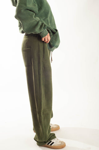 A person wearing olive green wide-leg sweatpants and a matching green sweatshirt, standing against a plain white studio background.