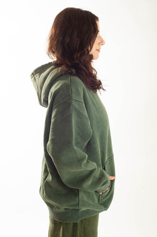 A woman shown from the side profile wearing an oversized sage green hooded sweatshirt and matching green pants against a plain white background.