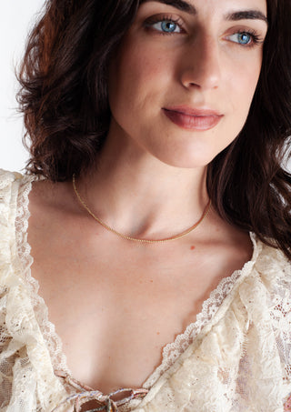 A close-up of a woman with dark brown hair wearing a delicate gold chain necklace and a cream lace-trimmed top against a plain white background.