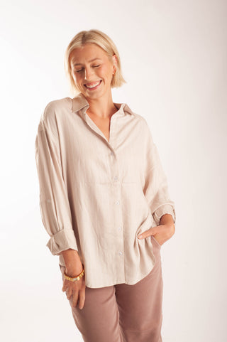 A smiling blonde woman wearing an oversized, light beige linen button-up shirt with rolled sleeves and brown pants, standing against a plain white studio background.