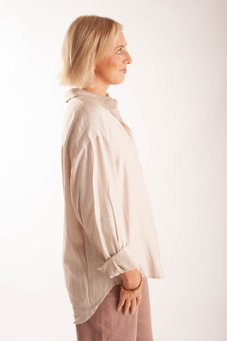 A woman in profile wearing a light beige linen button-up shirt and dusty pink trousers, standing against a plain white studio background.