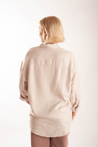 A woman with short blonde hair is shown from behind wearing a light beige linen button-up shirt and brown pants against a plain white studio background.