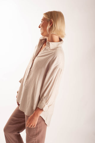 A woman in profile wearing a light beige, oversized long-sleeve button-down shirt and brown trousers, photographed against a clean white studio background.