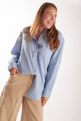 A woman wearing a light blue and white vertically striped button-down shirt and beige wide-leg pants, standing against a plain white studio background.