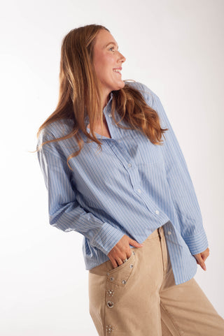 A woman wearing a light blue and white vertically striped button-down shirt and beige pants, posing against a plain white studio background.