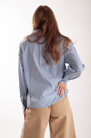 A woman shown from behind wearing a light blue and white vertically striped button-up shirt and beige pants, photographed against a plain white studio background.