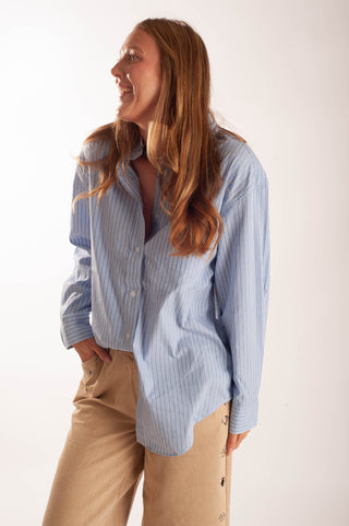 A woman wearing a light blue and white vertically striped button-down shirt and beige pants, standing against a plain off-white studio background.