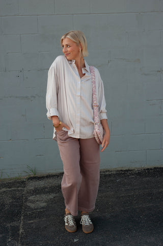 A woman stands against a gray painted brick wall wearing a loose white button-up shirt, dusty pink wide-leg pants, and brown sneakers, with a pink patterned shoulder bag.