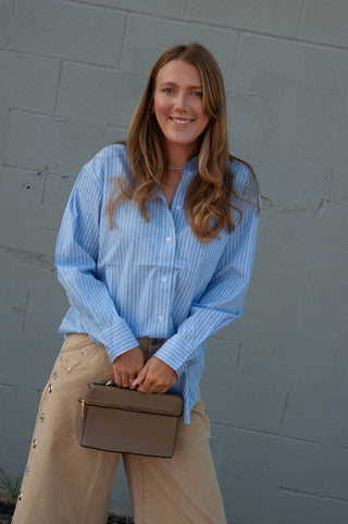 A woman stands against a gray painted brick wall wearing a light blue and white vertically striped button-down shirt, beige wide-leg pants, and holding a small taupe handbag.