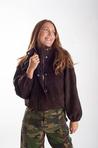 A woman wearing a dark brown suede-like zip-up jacket and green camouflage pants, standing against a plain white studio background.
