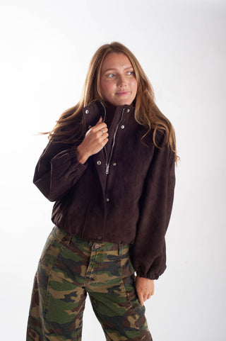A woman modeling a dark brown suede snap-button jacket with a high collar, paired with green and brown camouflage pants, against a plain white studio background.