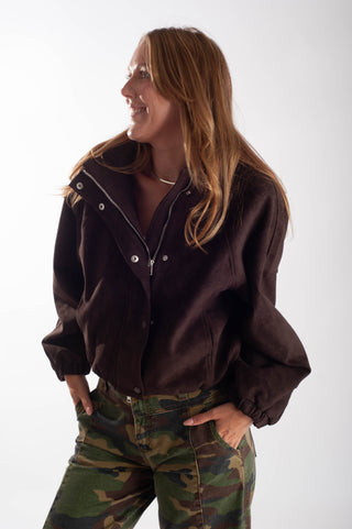 A woman wearing a dark brown cropped snap-button jacket with a high collar and a silver zipper, paired with green camouflage pants, posing against a light gray studio background.