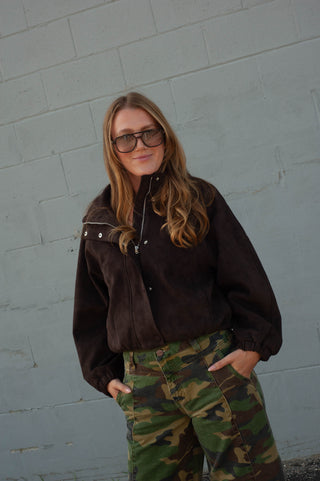 A woman wearing a dark brown suede zip-up jacket and green camouflage cargo pants, standing against a light gray painted brick wall.