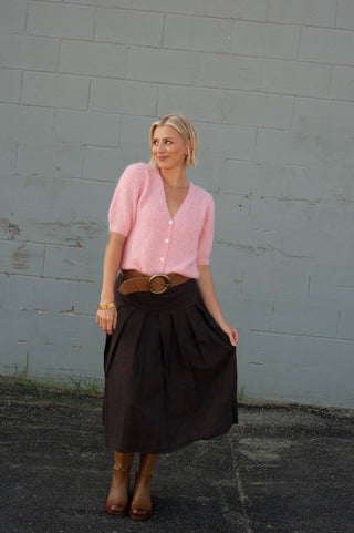 A woman stands outdoors against a gray concrete wall wearing a light pink short-sleeve knit cardigan, a dark brown pleated midi skirt, a wide brown belt with a large round buckle, and brown ankle boots.