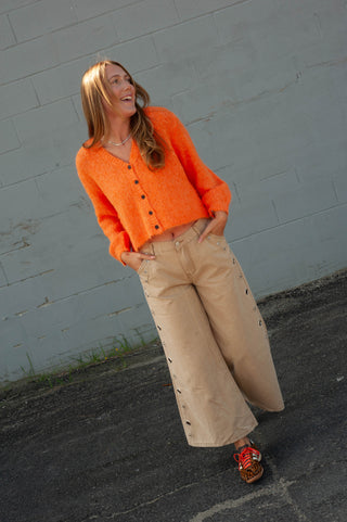 A woman wearing a bright orange fuzzy button-up cardigan, beige wide-leg pants, and red sneakers, standing outdoors against a gray brick wall.