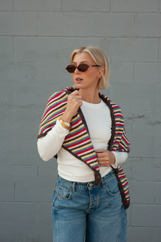 A woman wearing a colorful striped knit wrap shawl over a white long-sleeve top and blue jeans, standing against a gray brick wall.