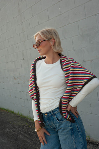 A woman wearing a white long-sleeve t-shirt, blue jeans, and a colorful striped knit poncho draped over her shoulders, standing against a light gray brick wall.