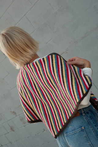 A person shown from behind wearing a colorful striped knit poncho or cape with blue jeans, standing against a gray brick wall.