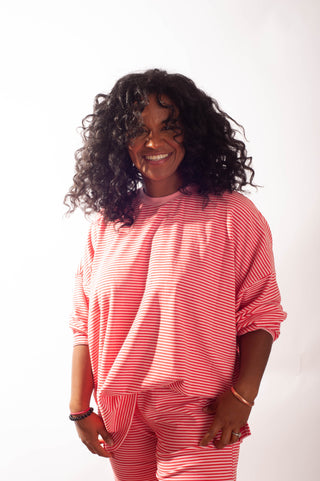 A smiling woman with curly black hair wearing a matching pink and white striped lounge set, consisting of a long-sleeve top and pants, against a plain white background.