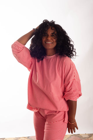 A woman with curly black hair wearing a matching pink and white striped lounge set, featuring a long-sleeve top and pants, posing against a plain white background.