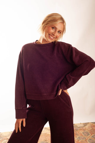 A woman modeling a matching deep plum corduroy lounge set, featuring a long-sleeve cropped sweatshirt and wide-leg pants, against a bright white studio background.