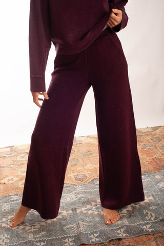 A person wearing a matching deep plum lounge set with a long-sleeve top and wide-leg pants, standing barefoot on a patterned rug against a plain light background.