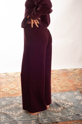 A person wearing dark red wide-leg pants and a matching dark top, standing barefoot on a patterned rug against a plain white wall.