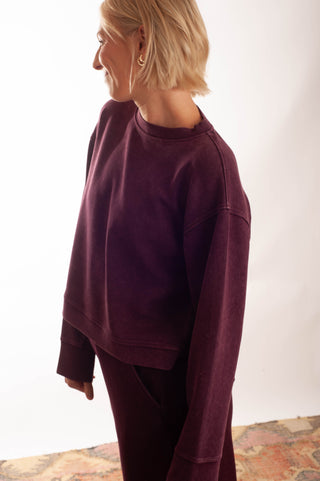 A woman wearing a dark plum crewneck sweatshirt and matching sweatpants, shown in profile against a plain white studio background.